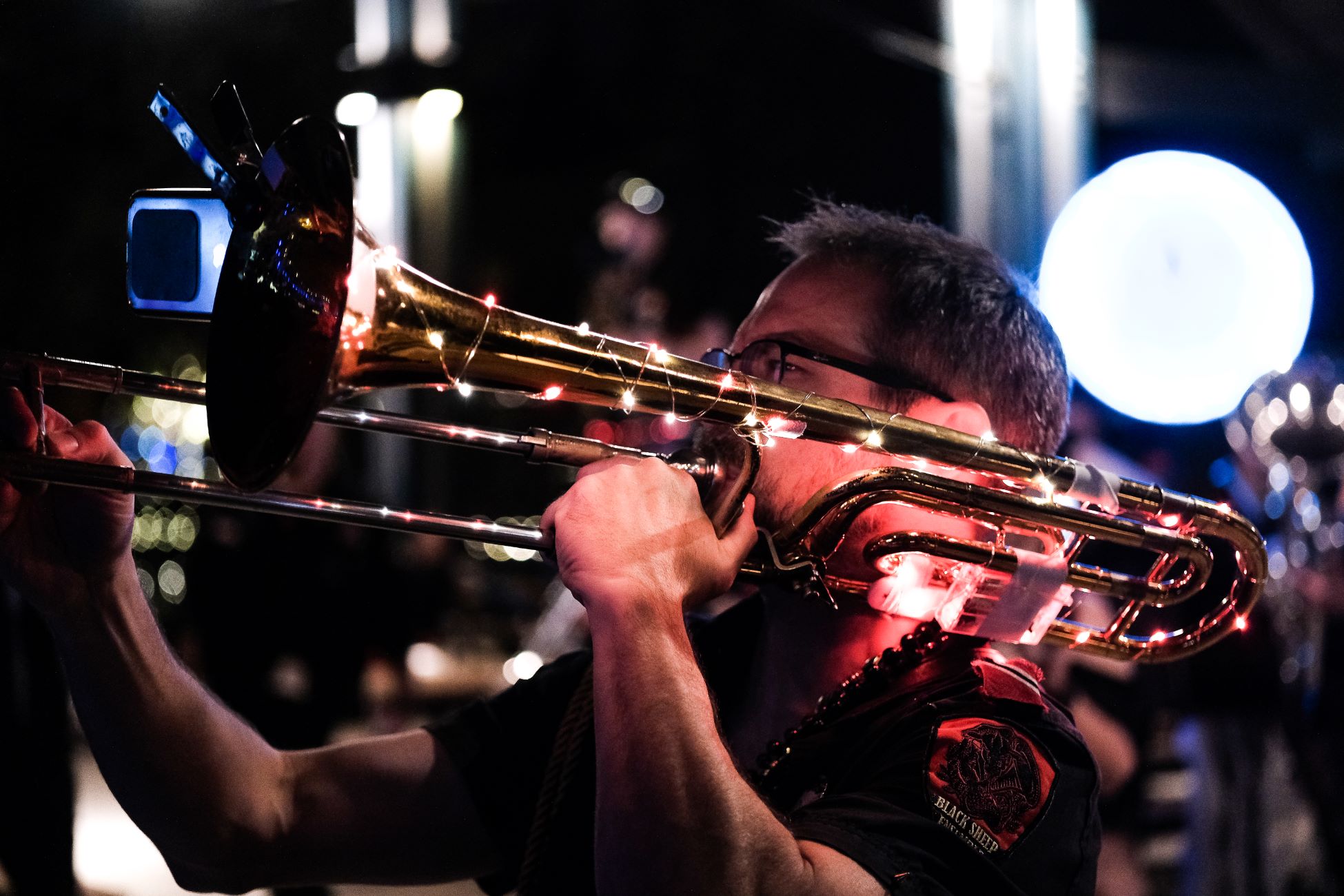 A trombone player plays music, as he marches in the Lantern Parade. His horn is covered in string lights. 