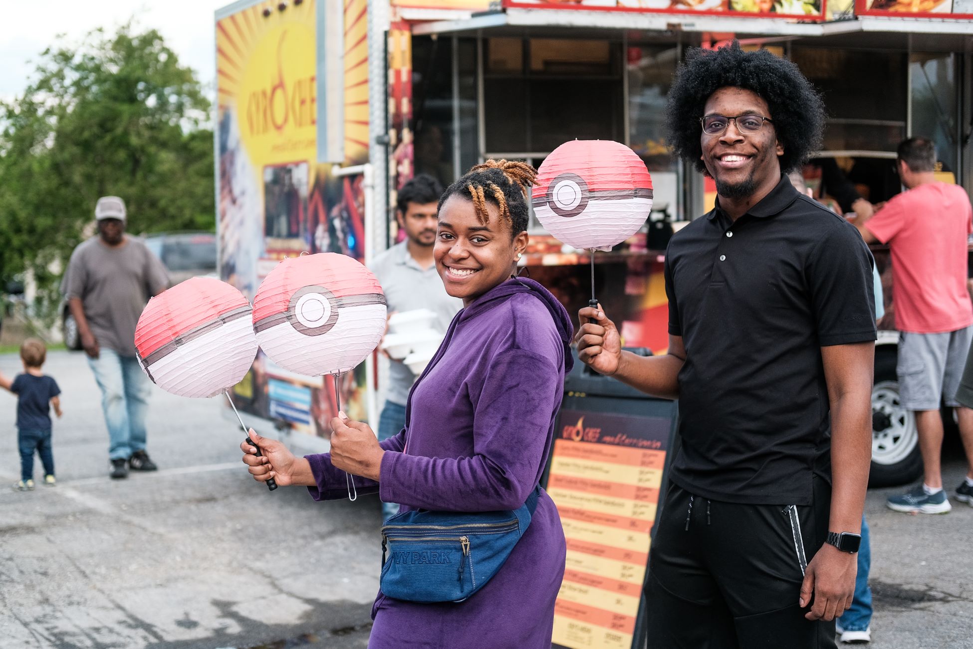 Two guests enjoy the Pre-Parade festivities. They are holding up lanterns that look like pokemon balls. In the background is a food truck.