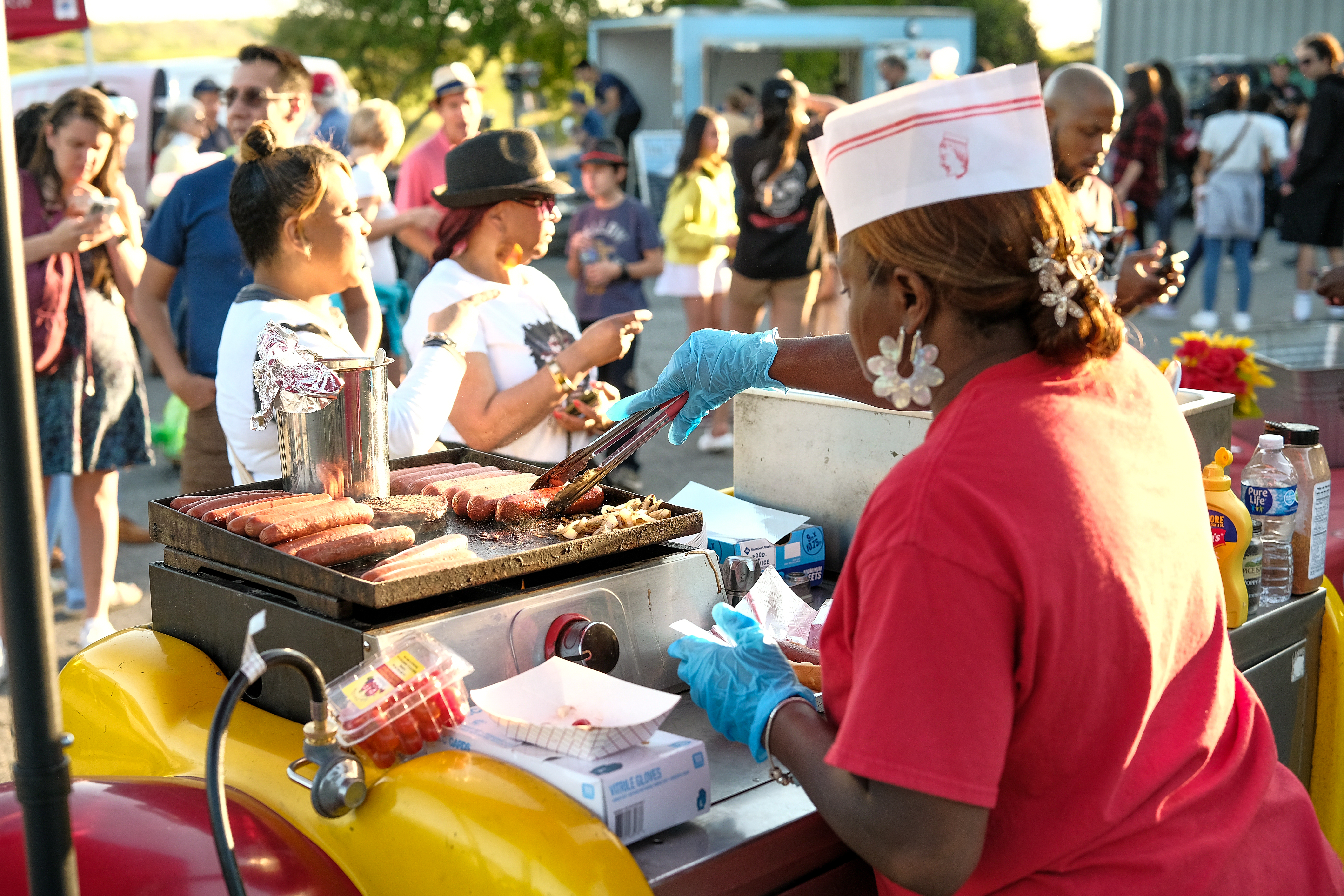 A food vendor, Dogs on Wheels, cooks hotdogs & sausages on a flat top grill. 